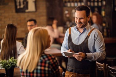 Happy waiter talking to female customer and writing her order on digital tablet in a bar. 