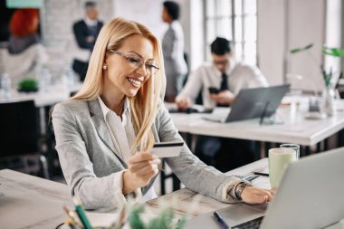 Happy businesswoman using laptop and credit card for online shopping while sitting at her desk in the office. 