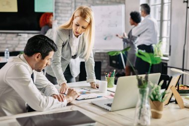 Mid adult businesswoman and her co-worker reading charts and analyzing business progress at work. There are people in the background. 