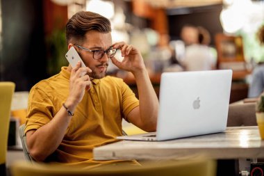 Pensive freelancer talking on cell phone while reading problematic e-mail on laptop in a cafe. 