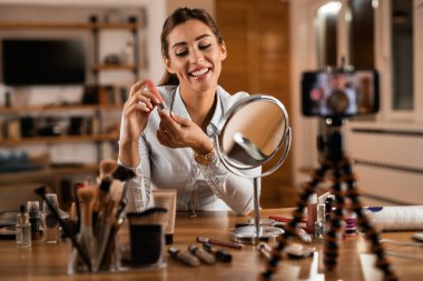 Young influencer holding lip balm while vlogging about make-up at home. 