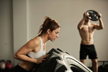 Strong athletic woman doing flip tire exercise during cross training. There is a man working out with weights in the background. 