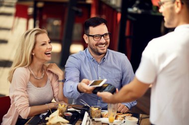 Happy couple having fun while making contactless payment via smart phone in a bar. 