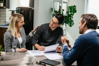 Happy couple communicating while singing a contract on a meeting with real estate agent. Focus is on young man. 