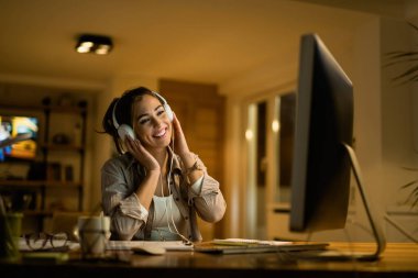 Young woman having fun at night while using desktop PC and listening music on headphones at home. 