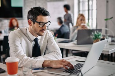 Young businessman sitting at office desk and working on laptop. There are people in the background. 