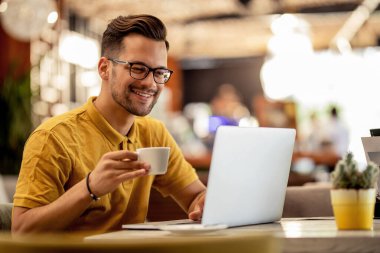Smiling man surfing the net on laptop while drinking coffee in a bar. 