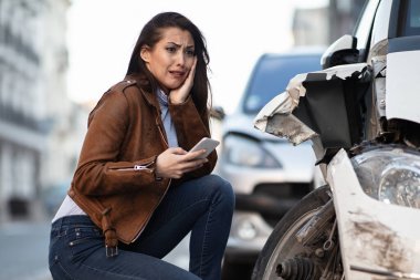 Young woman using a mobile phone while feeling shocked by car damage after an accident