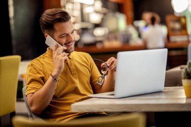 Young happy freelancer working on a computer while talking on smart phone in a bar. 
