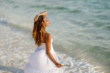 Young sensual woman in sundress standing at the shore with her eyes closed and enjoying in summer vacation. 