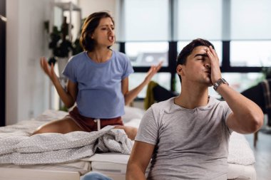 Young man feeling stressed out and having a headache while his girlfriend is arguing with him in the bedroom. 