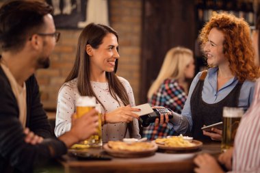 Happy waitress holding electronic reader while female customer is paying with smart phone via contactless payment. 