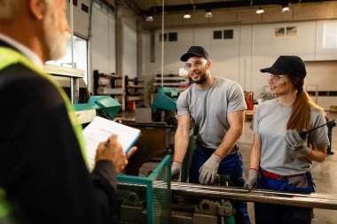 Two happy workers working at the machine and communicating with senior businessman who is overseeing them in industrial building. 
