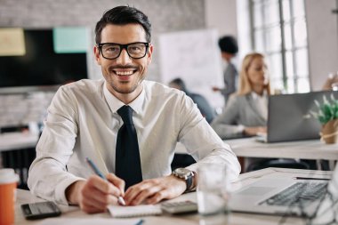 Young smiling financial advisor working on paperwork and looking at camera in the office. There are people in the background. 