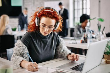 Redhead businesswoman using laptop and writing notes in her notepad while listening music on headphones at work. 