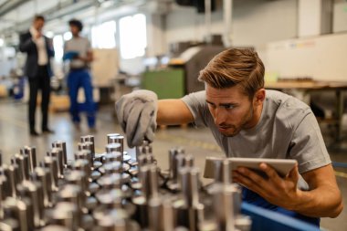 Young steel worker examining manufactured rod cylinder while using digital tablet in distribution warehouse. 
