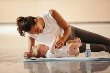 Happy athletic mother playing with her baby boy who is lying on exercise mat in health club. 