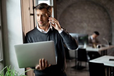 Businessman holding laptop and talking on mobile phone while standing by the window in the office. 