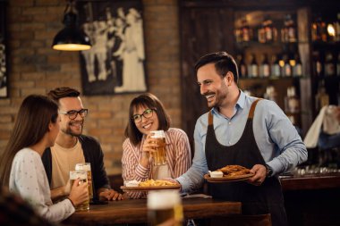 Happy waiter giving food to group of guests while they are drinking beer in a pub. 
