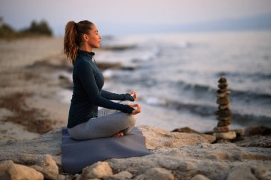 Side view of Zen-like woman with eyes closed practicing Yoga on a rock by the sea. 