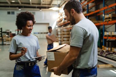 Happy black female worker going through check list and talking to her coworker who is carrying cardboard box in distribution warehouse. 