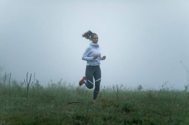 Full length of athletic woman jogging through the filed on cold misty weather. Copy space. 