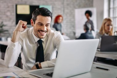 Young happy businessman using computer in the office and celebrating good news. There are people in the background. 