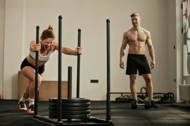 Young sportswoman doing sled push exercise and shouting during sports training in a gym while her friend is watching her. 