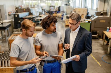 Quality control inspector and young workers communicating while using digital tablet and going through reports in steel factory. 
