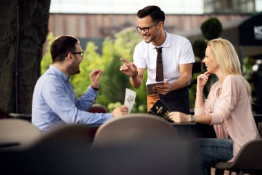 Happy waiter serving a couple and using touchpad while taking their order in a cafe. 