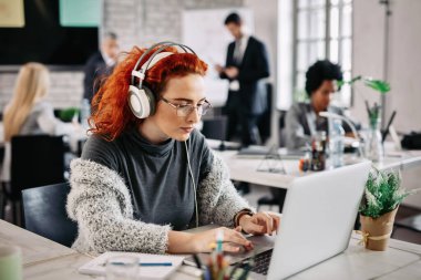 Young businesswoman surfing the net on laptop while listening music on headphones and working in the office. There are people in the background. 