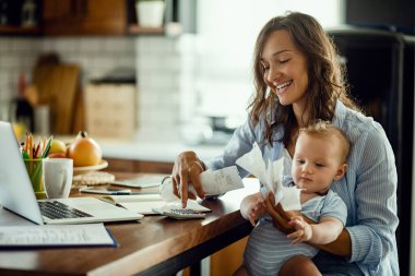 Young working mother with baby using calculator and going through bills while calculating home finances. 