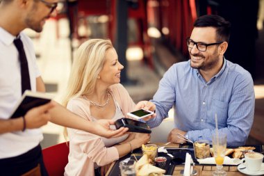 Happy couple making contactless payment with smart phone after lunch in a restaurant. 