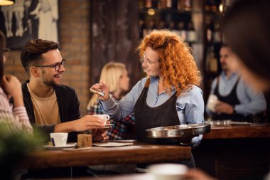 Happy waitress pouring cinnamon in coffee cup while serving young man in coffee shop. 