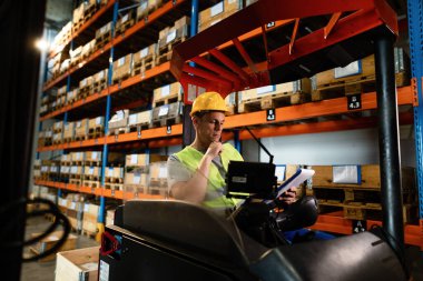 Manual worker sitting in a forklift and reading paperwork while working in warehouse. 