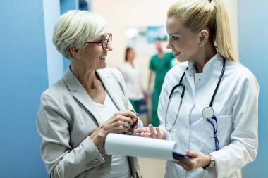 Happy female doctor and mature woman talking while analyzing medical reports in the hospital. 