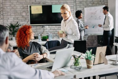 Happy businesswoman talking to her female colleague during coffee break in the office. There are people in the background. 