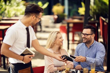Happy man making contactless payment with smart phone while being with his girlfriend in a restaurant. 