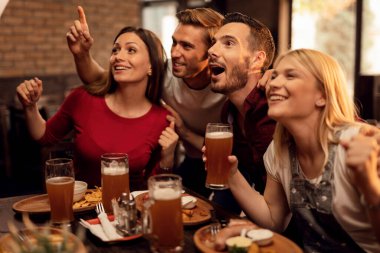 Group of excited friends cheering for their team and having fun while watching sports game and drinking beer in a bar.