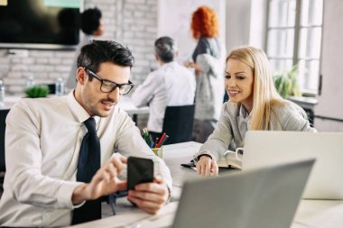 Mid adult businesswoman smiling while her male colleague is showing something on smart phone to her. 