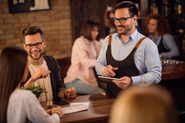 Happy waiter with touchpad communicating with customers and taking their order in a pub. 