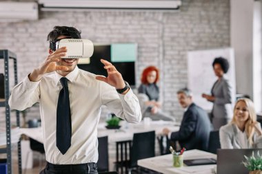 Playful businessman using virtual reality simulator and having fun at work. There are people in the background. 