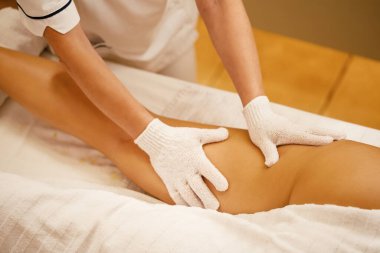 Close-up of therapist with exfoliating gloves massaging woman's leg during spa treatment. 