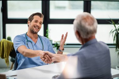 Happy doctor shaking hands with senior patient while talking to him during consultations at clinic.