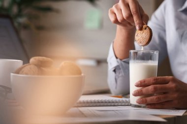 Close up of businesswoman having a snack at work and dipping biscuit in a glass of milk. 