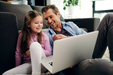 Happy little girl and her father watching something on a computer ad having fun together at home. 