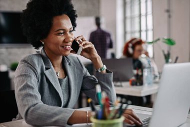 Happy African American businesswoman working on a computer and communicating on mobile phone in the office. There are people in the background.