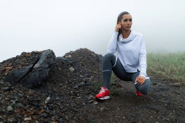 Young sportswoman taking a break after exercising and listening music over earphones in nature against foggy sky. Copy space. 