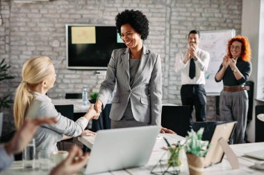 Happy African American businesswoman shaking hands with her female colleague in the office while other co-workers are applauding them. 