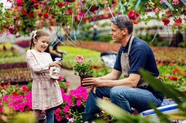 Happy little girl having fun with her father while using spray bottle and watering potted flowers at plant nursery. 